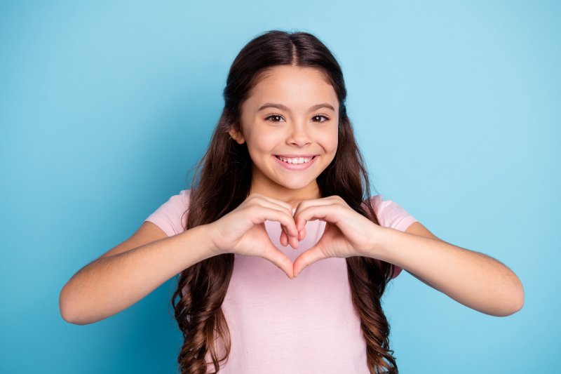 a young girl wearing a pink blouse and making a heart with her hands smiles